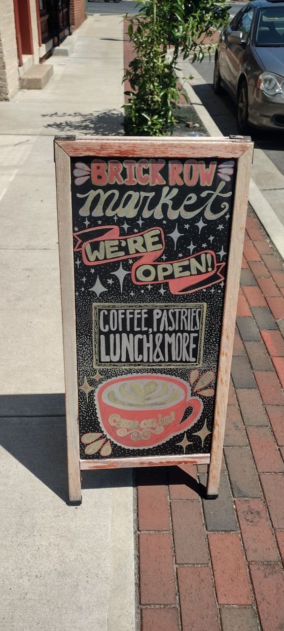 Hand-painted A-frame sandwich board reading Brick Row Market We're Open · Coffee, Pastries, Lunch &amp; More · Come on in