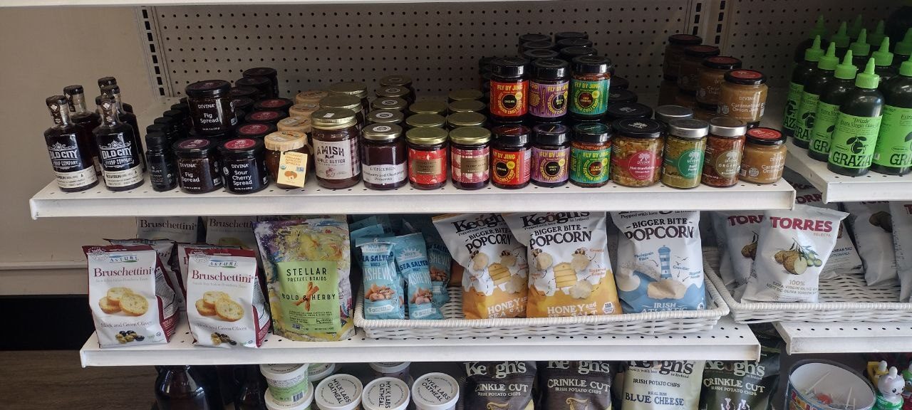 Market shelf with Old City flavor syrups, Divina fig spread, Amish maple butter, jarred preserves, Fly By Jing chili, Stellar pretzels, Keogh's popcorn, and Graza olive oil