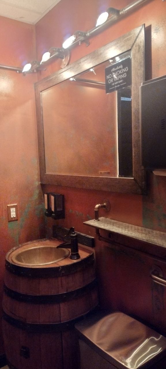 Cafe bathroom with a copper sink basin set into a whiskey barrel, patinated orange-plaster walls, a wide bronze-framed mirror, and industrial pipe vanity lighting