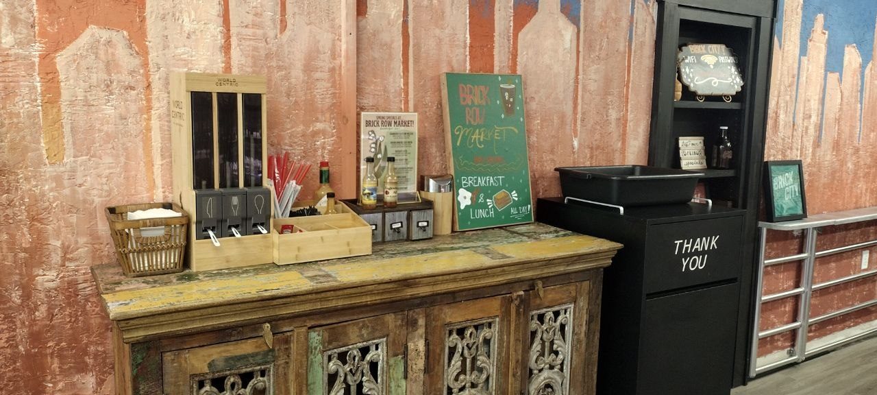 Vintage wooden console with self-serve coffee station — carafes, sugar caddy, straws, syrups — and a green Brick Row Market chalkboard reading Breakfast &amp; Lunch All Day, with a Thank You bin alongside against a painted rust-red wall