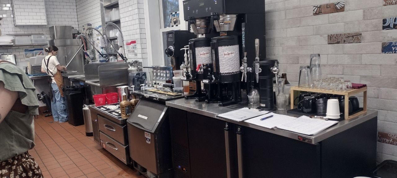 Twin batch coffee brewers and stacked glassware on the back-of-house bar, staff working the dish station behind, multi-color subway-tile accents