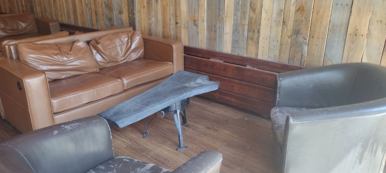 Lounge area with tan leather couches and a leather armchair around an industrial slate-top coffee table, against a pallet-wood wall