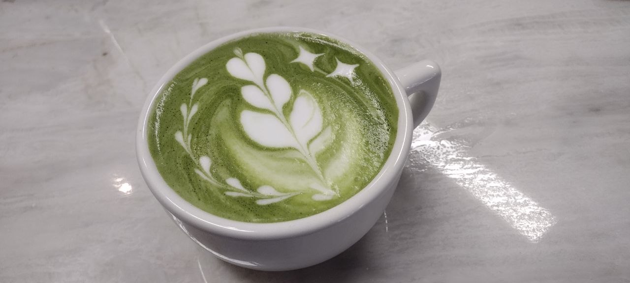 The flagship matcha latte at Brick Row Market — emerald green with an elaborate fern and starburst leaf-art pour on marble