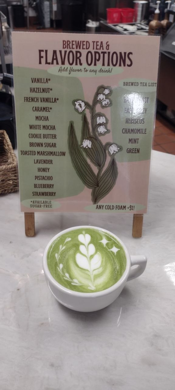 Matcha latte beside the hand-painted Brewed Tea & Flavor Options menu board listing 14 flavors and 6 brewed teas