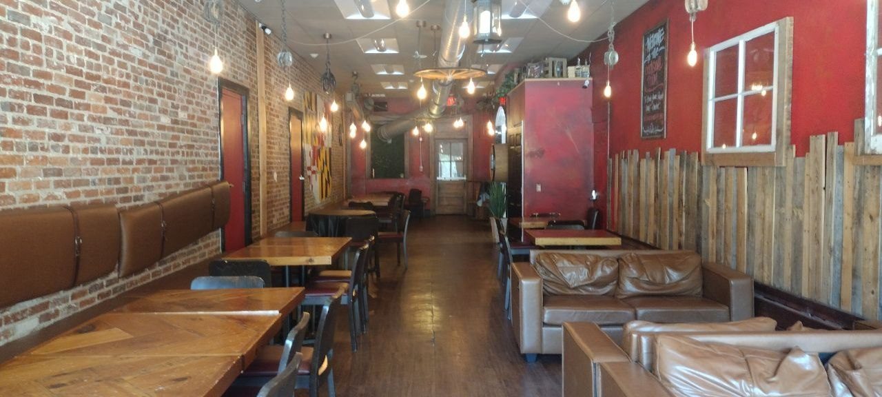 Wide interior shot of the dining room — exposed brick on the left with banquette seating, leather couches on the right, pallet-wood wainscoting, Maryland flag mural on the far wall, Edison bulbs throughout