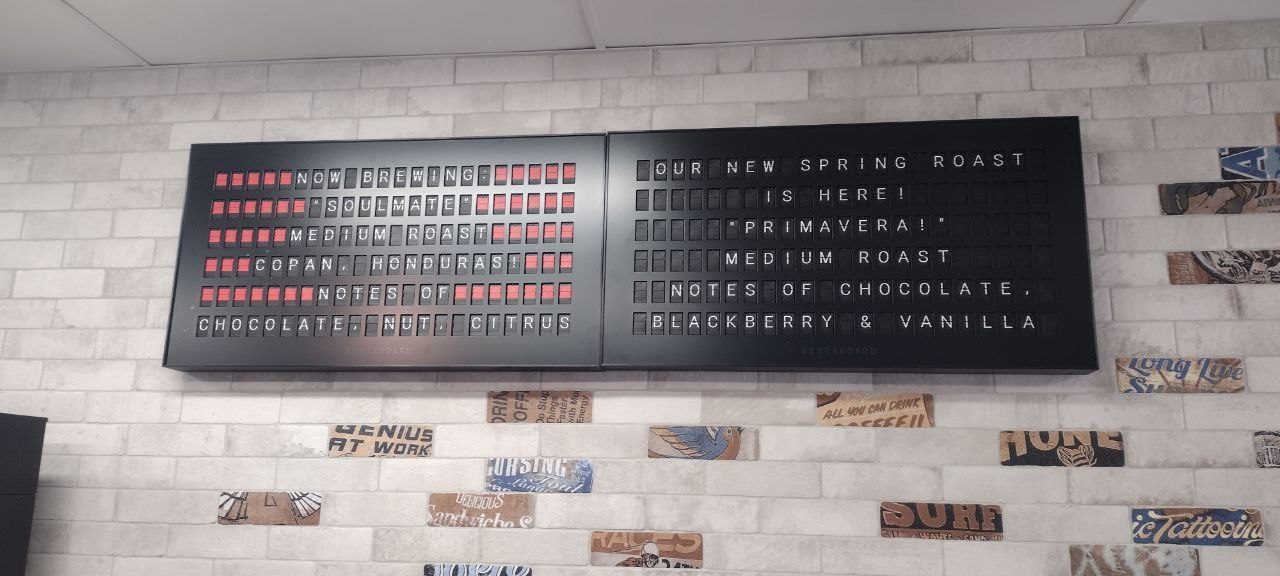 Twin Vestaboard flip-letter displays on the brick wall — left panel reads Now Brewing Soulmate Medium Roast Copan Honduras notes of chocolate nut citrus; right panel reads Our New Spring Roast Is Here Primavera Medium Roast notes of chocolate blackberry &amp; vanilla