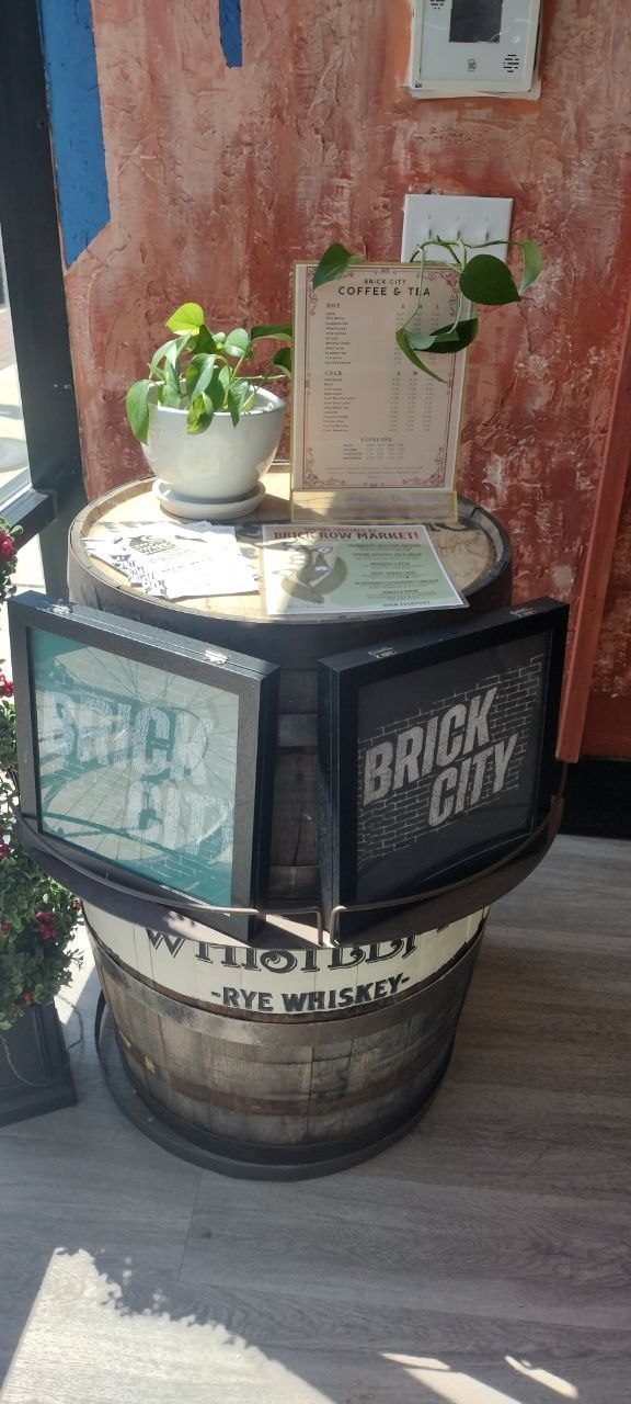 Rye whiskey barrel by the window topped with the Brick City Coffee &amp; Tea menu and a potted pothos, framed Brick City prints leaning against the front
