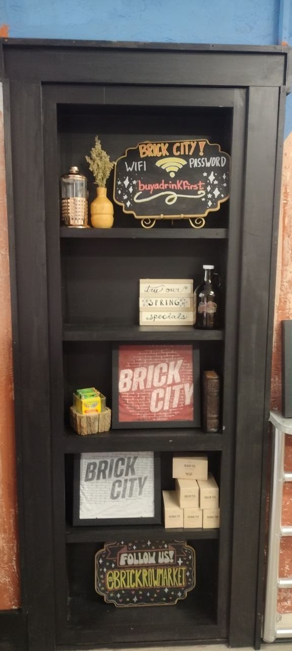 Black bookshelf with the hand-painted Brick City Wi-Fi password sign (buyadrinkfirst), Try Our Spring Specials sign, framed Brick City prints, tea boxes, and a Follow Us @brickrowmarket sign on the bottom shelf